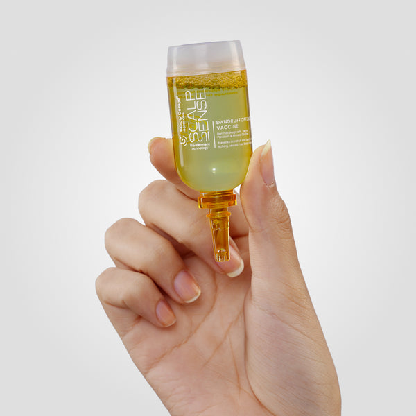 A hand holds a container of yellow liquid labeled "ScalpSense Dandruff Defense Vaccine" against a plain background.