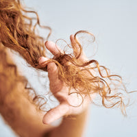 A close-up of a hand addressing a hair concern by holding a section of curly, light brown hair against a plain background.